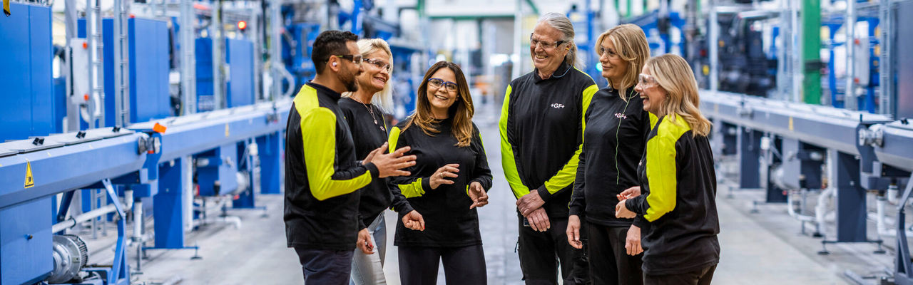 Six GF employees stand together in a modern industrial facility, engaged in a group discussion.