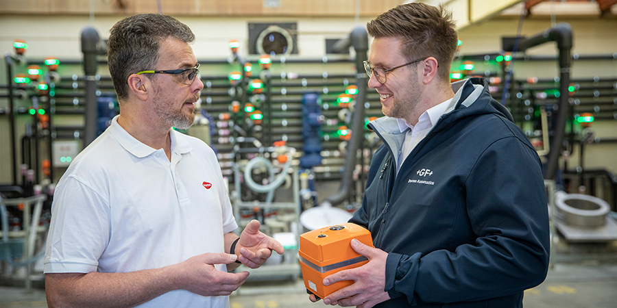 two men speaking to each other in front of a process automation installation. one men holding up an actuator